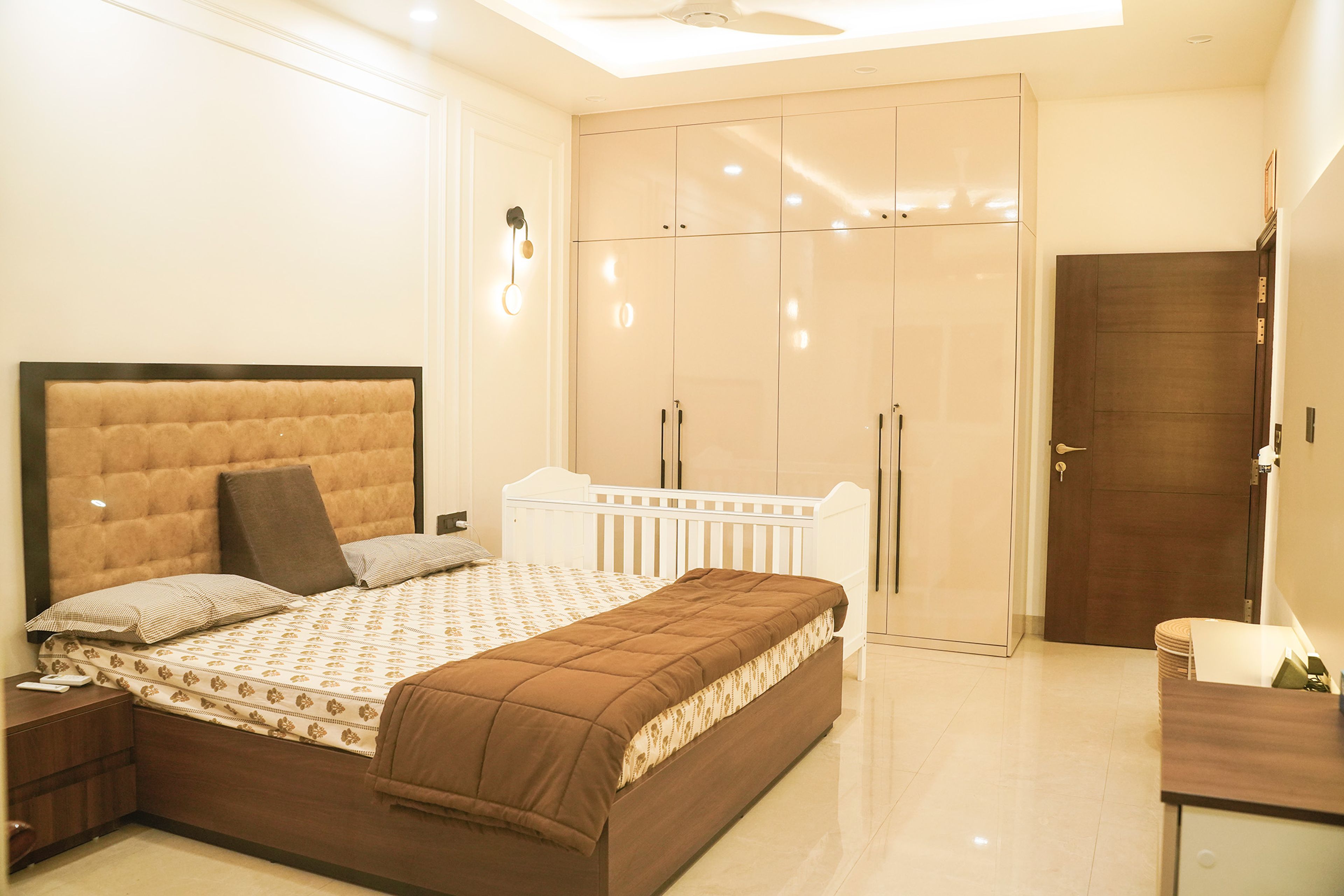 Master Bedroom Design with Beige Upholstered Bed, 4-Door Glossy Wardrobe, and White Baby Cot