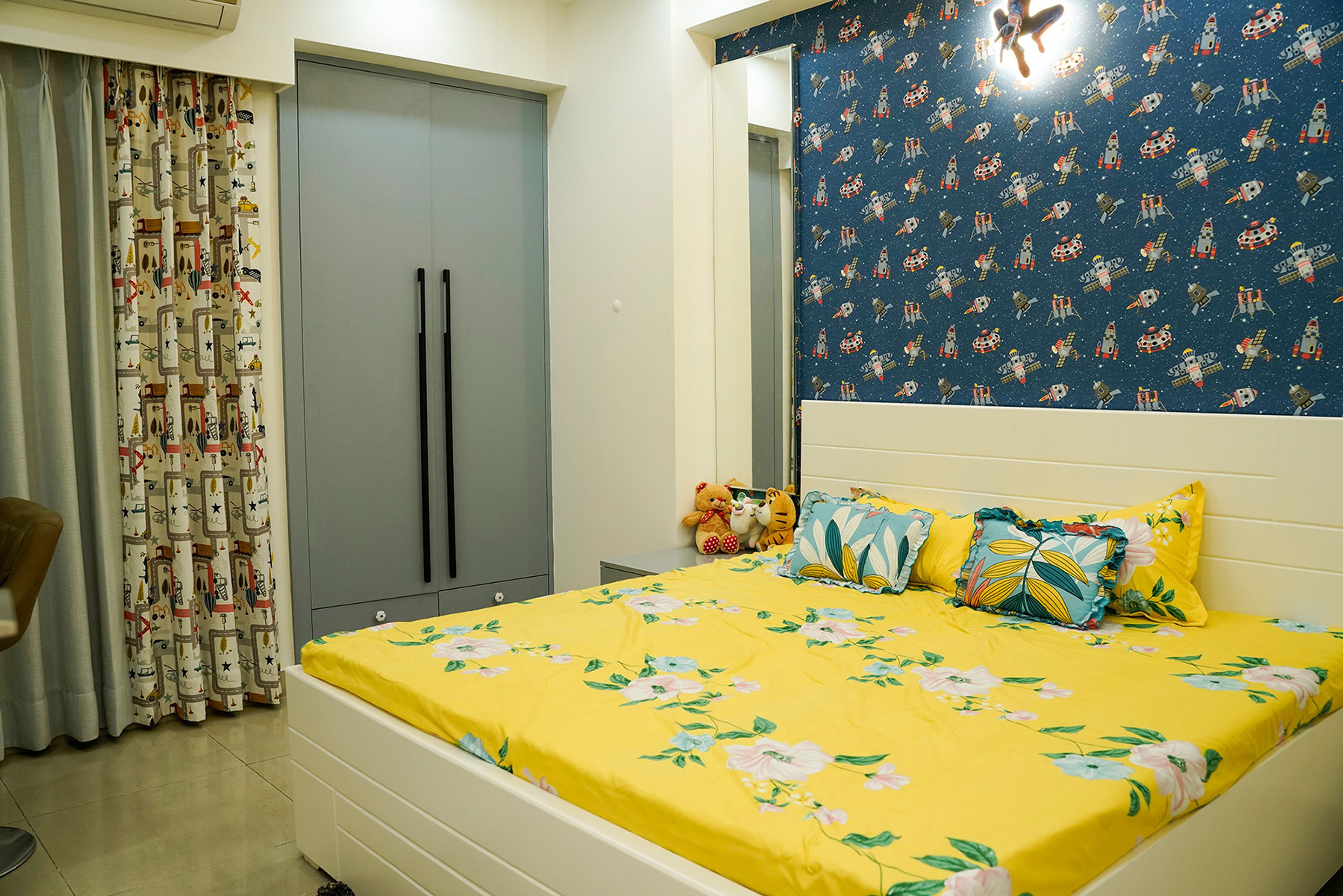 Space-themed kids’ bedroom with blue accent wall, yellow floral bedding, and sleek grey wardrobe
