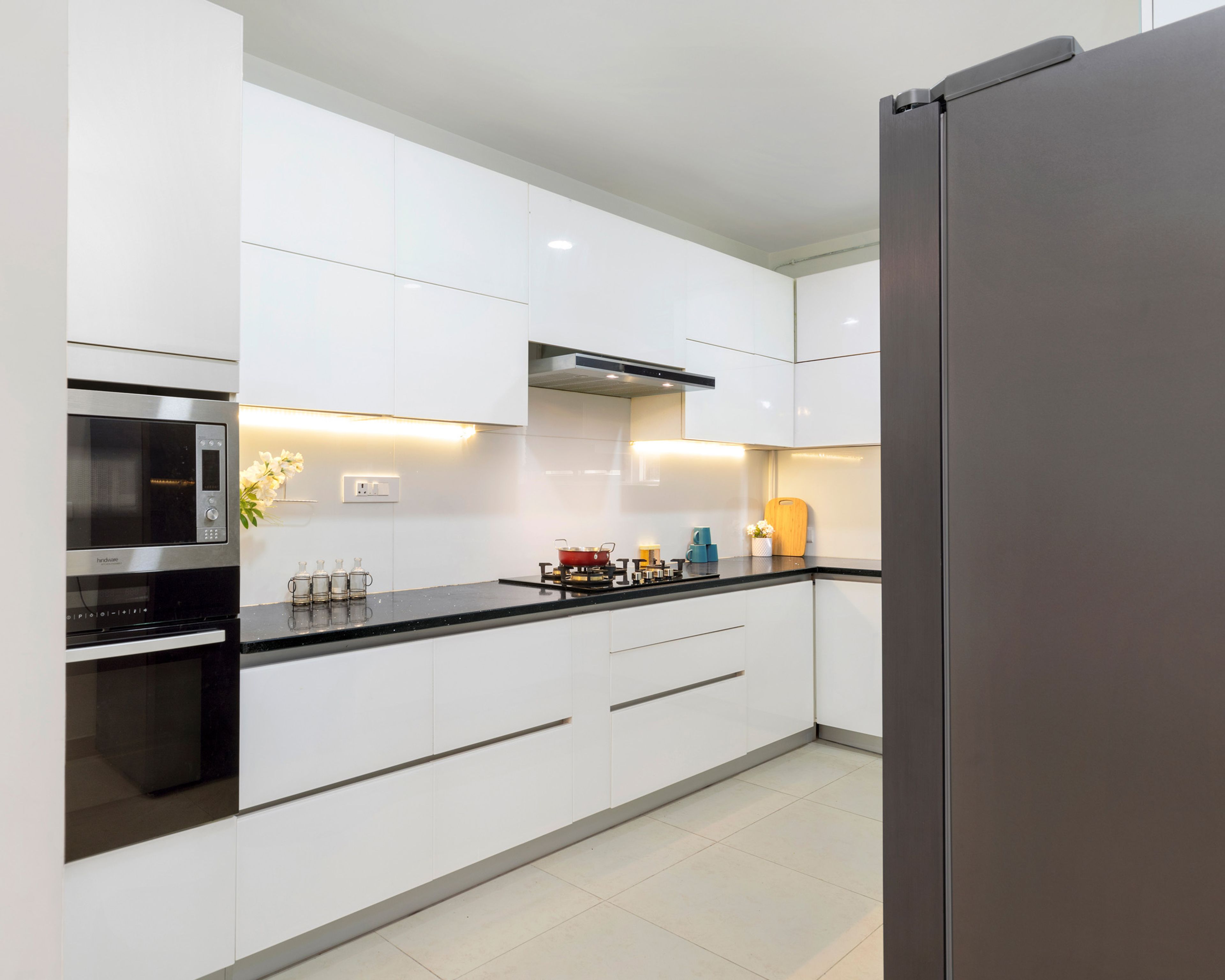 L-shape white glossy kitchen design with black kitchen countertop and under-cabinet lighting - Livspace