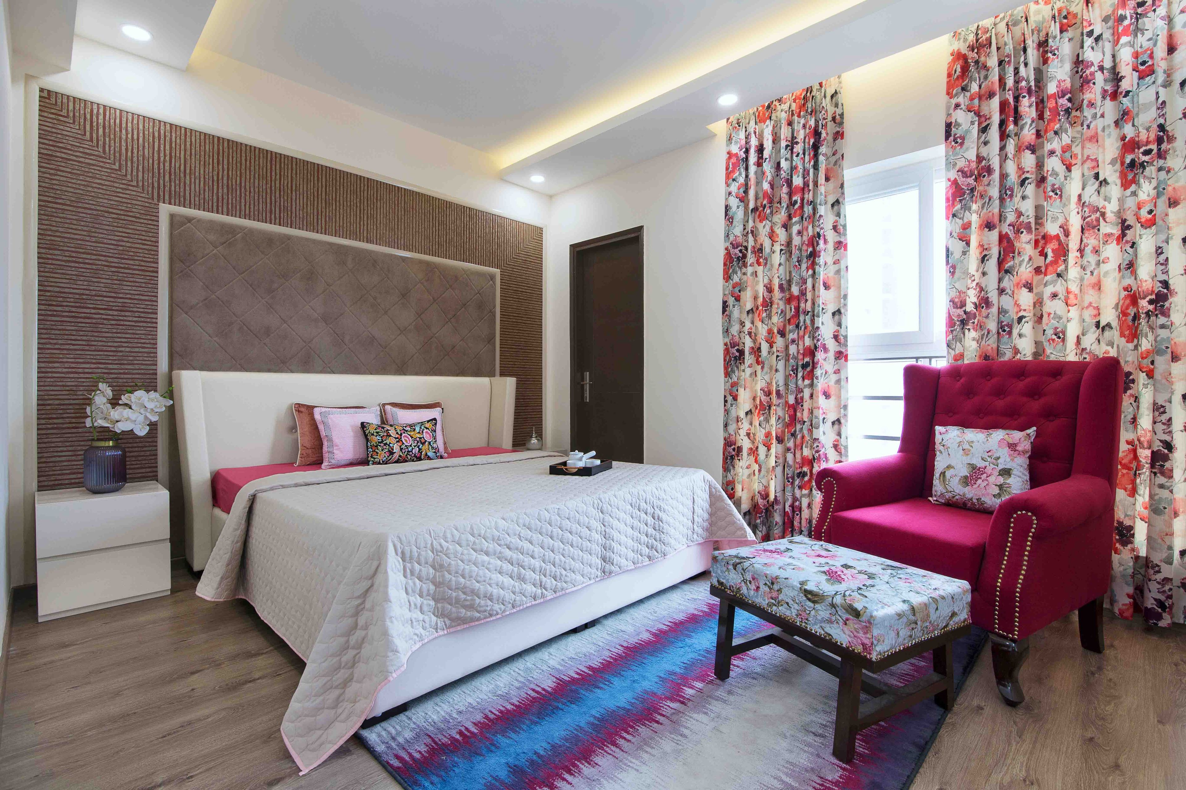 Master Bedroom With Large Brown Tufted Wall Panel, Off-White Headboard And Magenta Accent Chair With Floral Footstool - Livspace