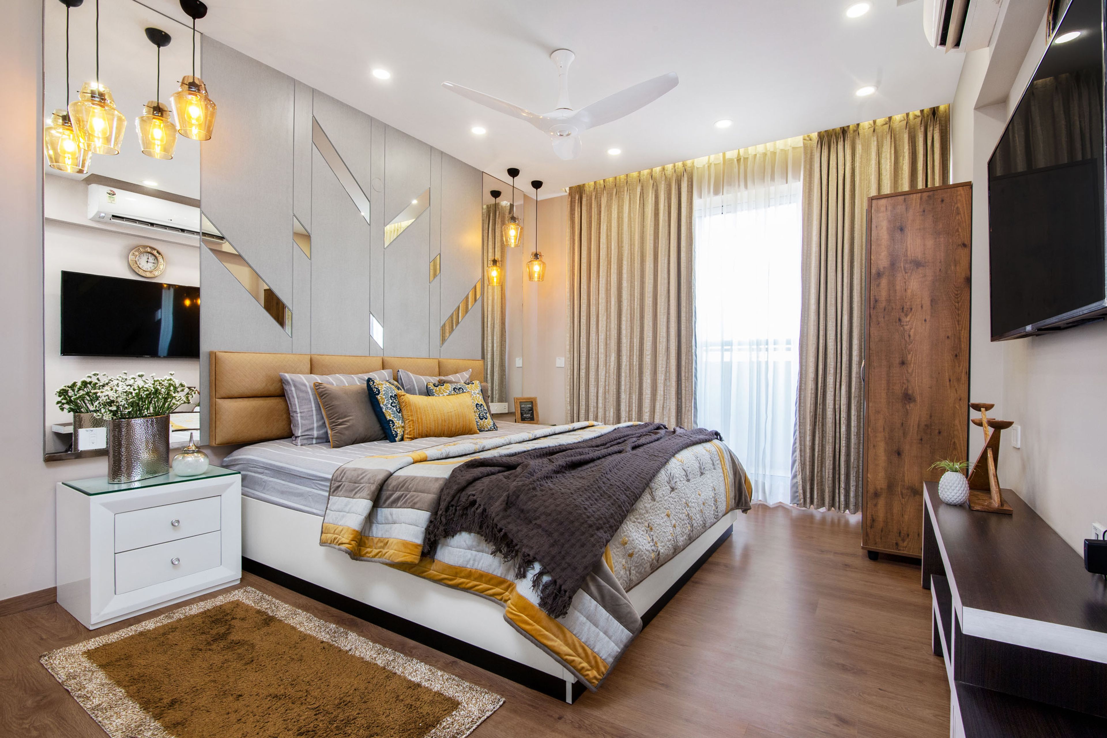 Spacious Master Bedroom With White Bed And Amber Headboard, Silver Accent Wall With Mirrored Panels And Wood And White TV Unit- Livspace