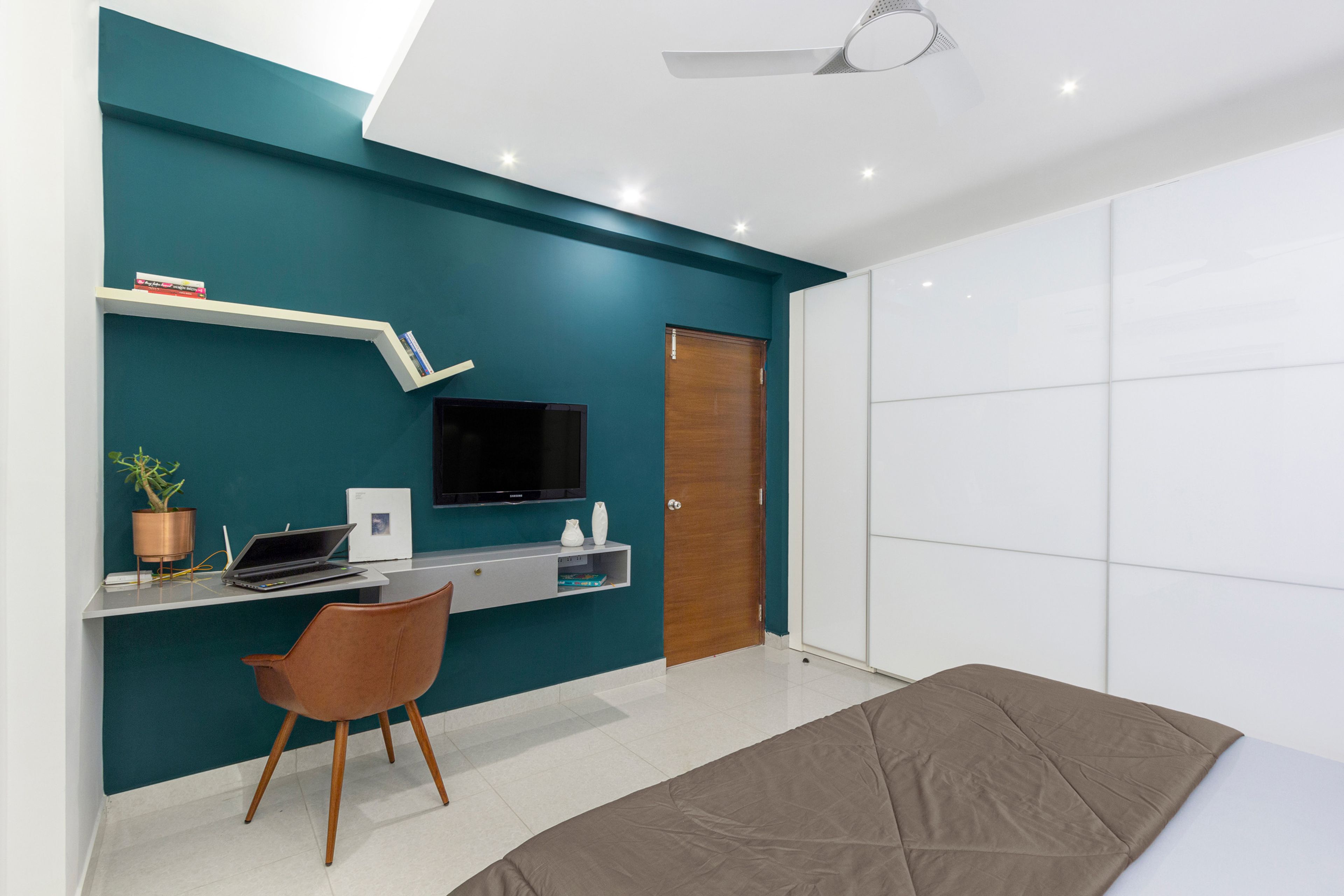 Wall-mounted grey TV unit in master bedroom and integrated study unit against teal blue accent wall - Livspace