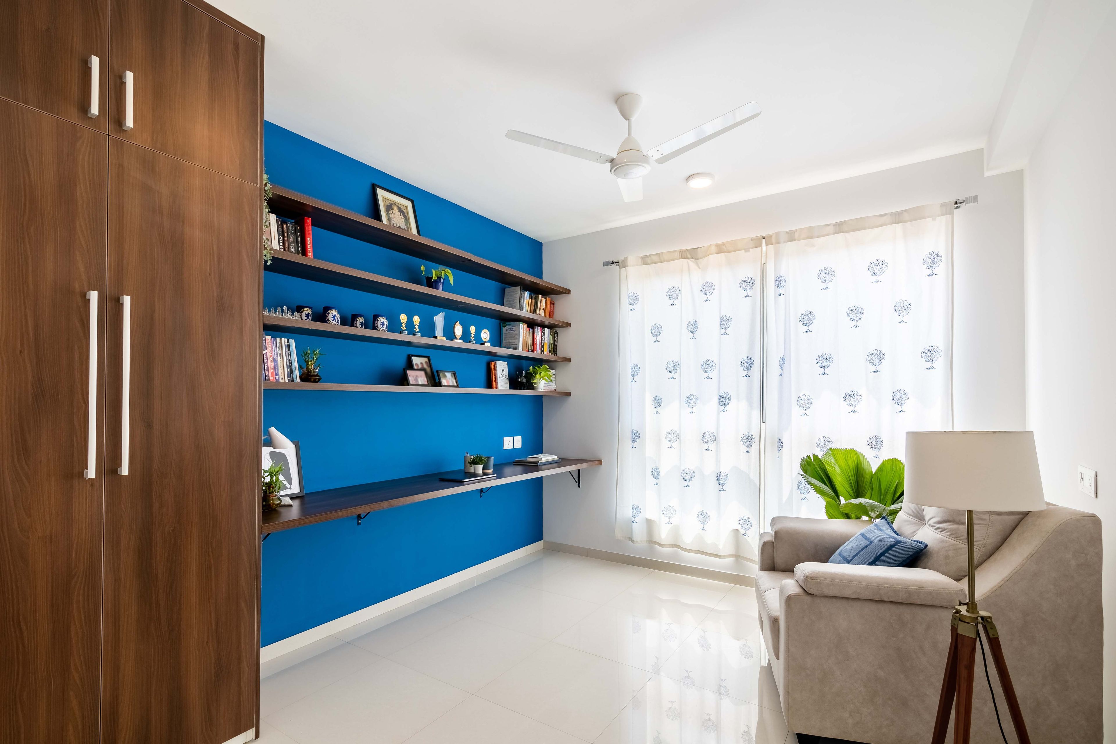 Home office design with bright blue accent wall, wooden shelves,  2-door wooden swing wardrobe and a beige sofa - Livspace